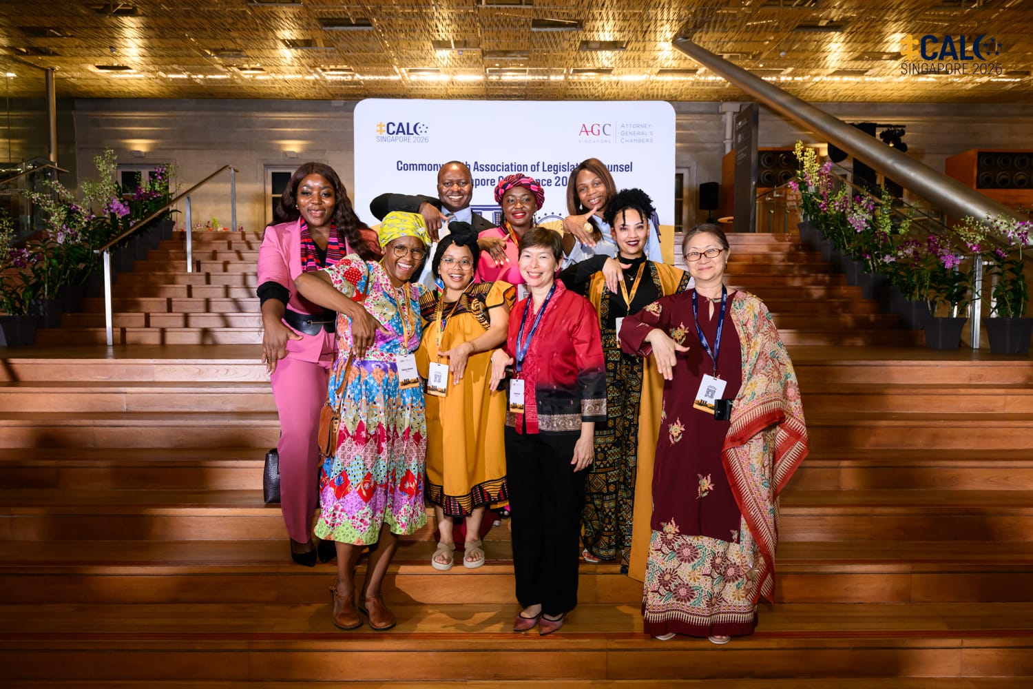A diverse group of conference delegates, including Ms Jeanne Lee, Chief Legislative Counsel of Singapore, in colourful traditional attire pose together on wooden steps in front of the Commonwealth Association of Legislative Counsel Singapore Conference 2026 backdrop.